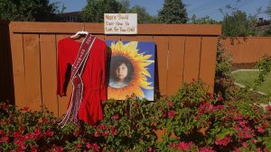 Portrait of Ashlynne Mike next to ceremonial dress, and a handwritten sign that reads "No one should have to look for their child."